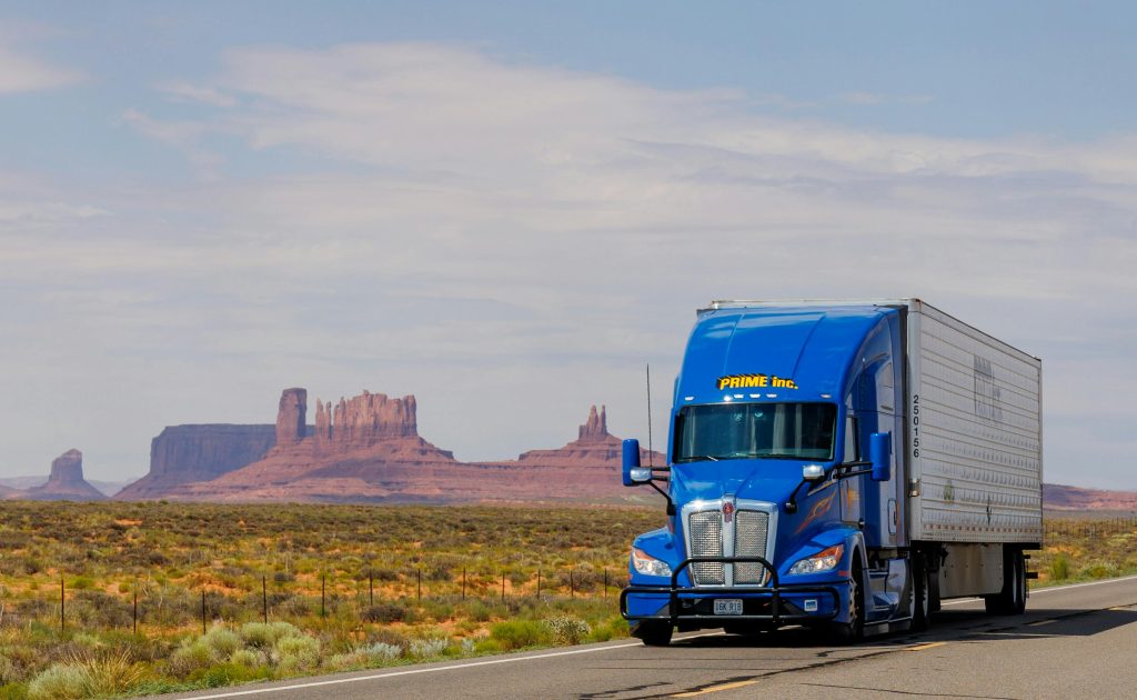 A blue truck travels on the highway with Monument Valley's iconic mesas in the background.