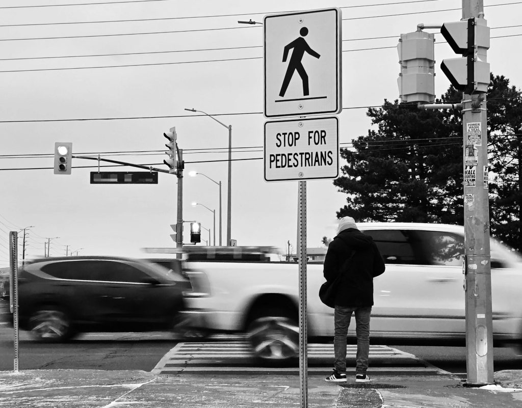 Black and white photo of a pedestrian waiting at a crosswalk in Canada.