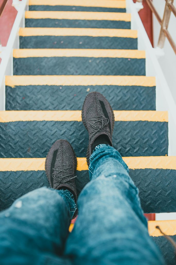 A person's feet in sneakers taking a step down outdoor stairs with yellow stripes.